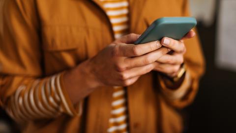 Nahaufnahme einer Hand, die ein Smartphone hält. Die dazugehörige Person trägt ein orangeweiß gestreiftes Shirt und eine orangefarbene Jacke.