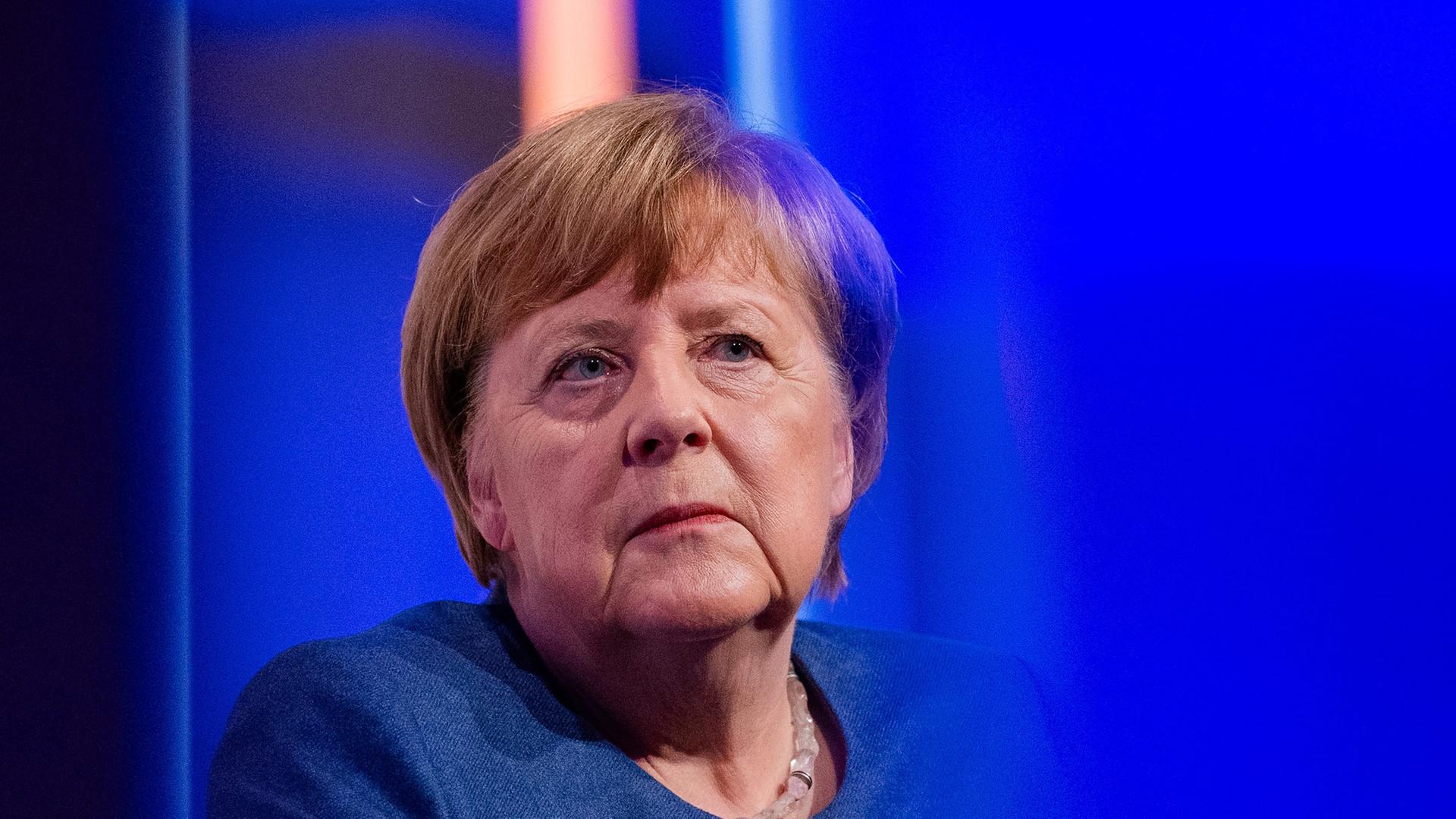 Angela Merkel (CDU) sitzt auf einer Bühne mit blauem Hintergrund und trägt eine blaues Sakko.
