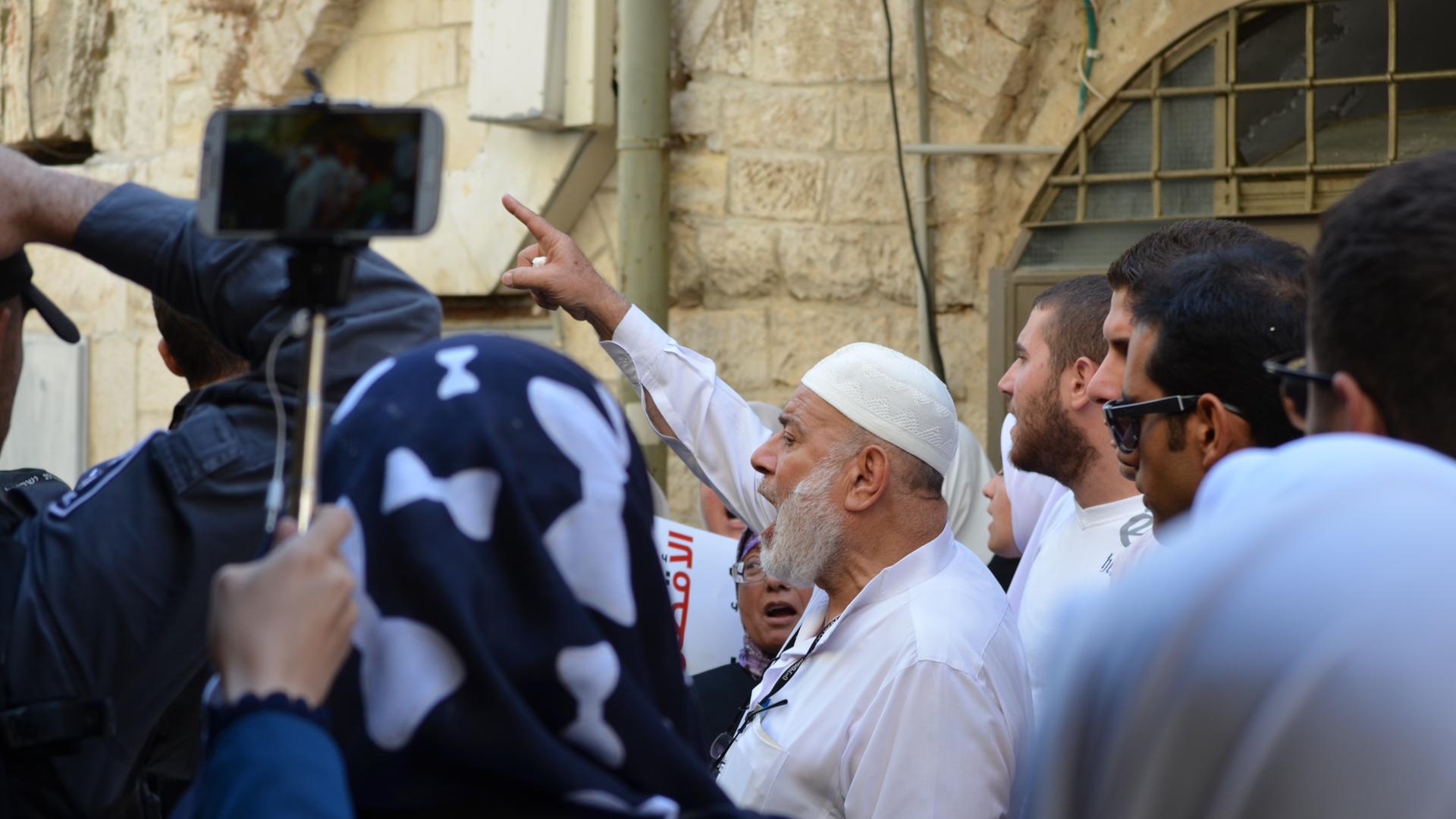 Muslimische Männer, teils mit erhobener Faust vor einer sandfarbenen Wand in Jerusalem während einer Demonstration. Eine Person filmt die Gruppe mit einem Smartphone.