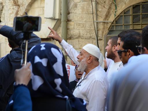Muslimische Männer, teils mit erhobener Faust vor einer sandfarbenen Wand in Jerusalem während einer Demonstration. Eine Person filmt die Gruppe mit einem Smartphone.