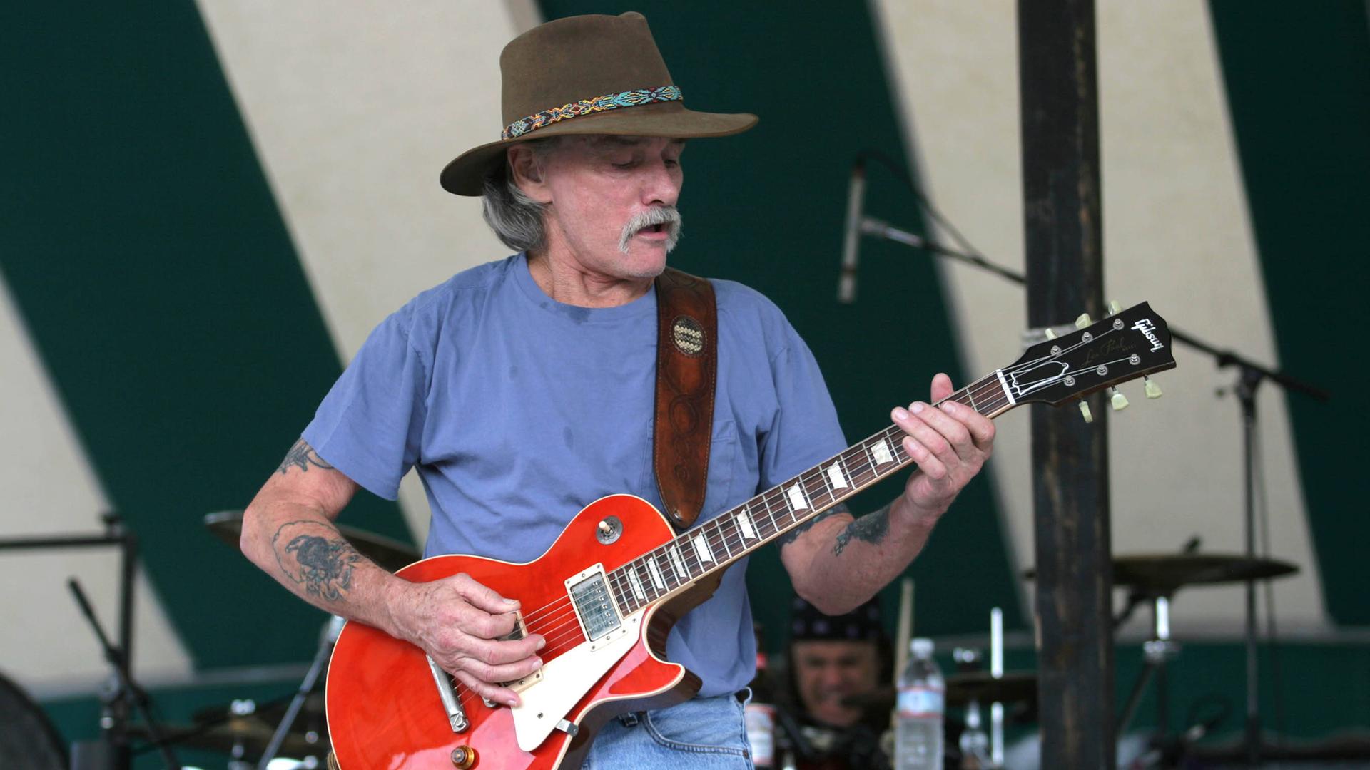 Dickey Betts trägt einen brauen Cowboy-Hut und steht damit auf einer Outdoor-Bühne mit Mikrofonen, während er auf seiner roten Gitarre spielt und dabei singt.