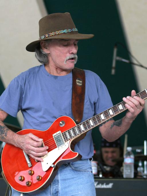 Dickey Betts trägt einen brauen Cowboy-Hut und steht damit auf einer Outdoor-Bühne mit Mikrofonen, während er auf seiner roten Gitarre spielt und dabei singt.