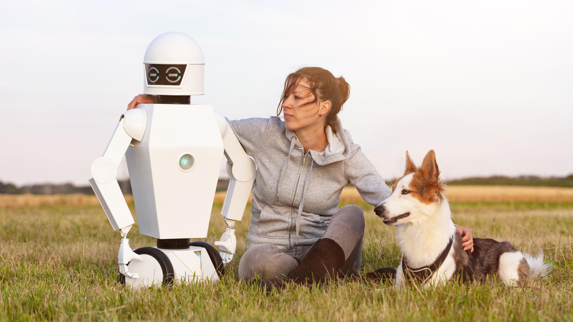 Eine Frau sitzt mit einem Hund und einem Roboter auf einer WIese.