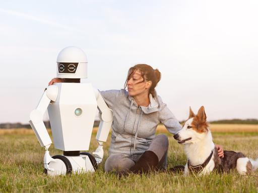 Eine Frau sitzt mit einem Hund und einem Roboter auf einer WIese.