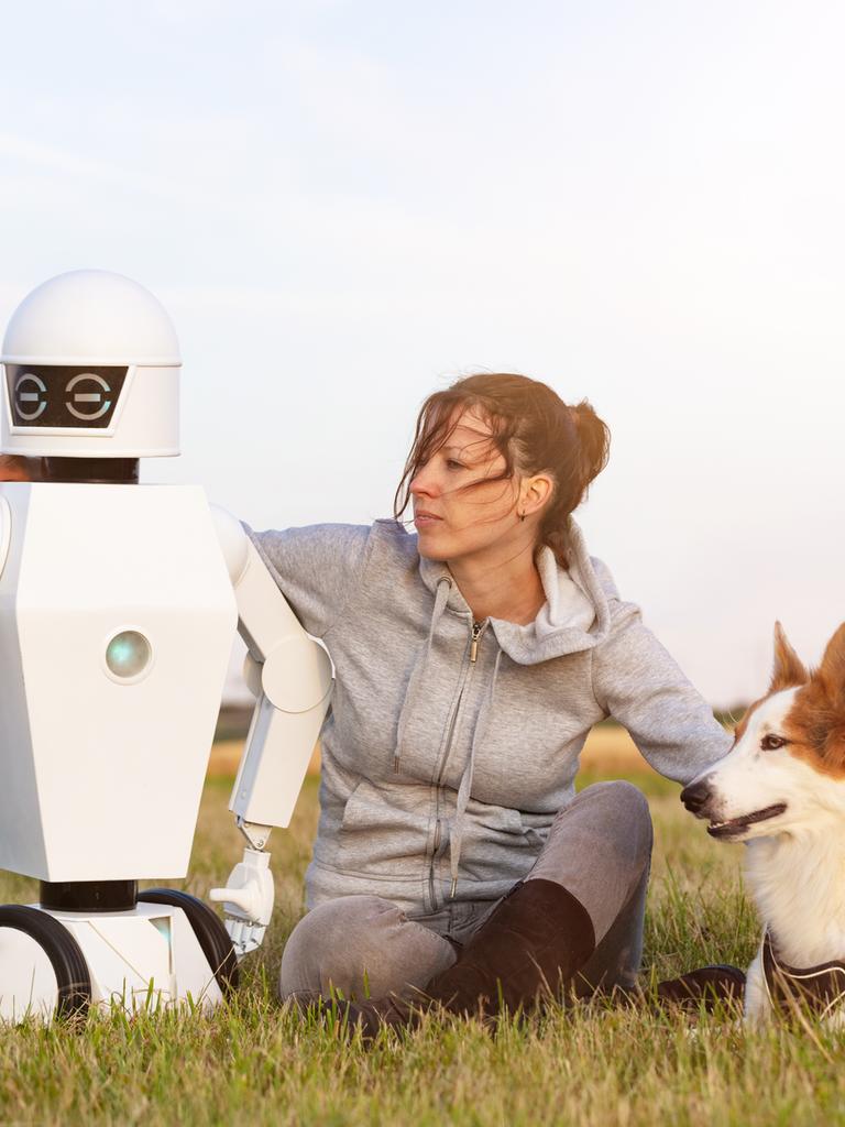 Eine Frau sitzt mit einem Hund und einem Roboter auf einer WIese.