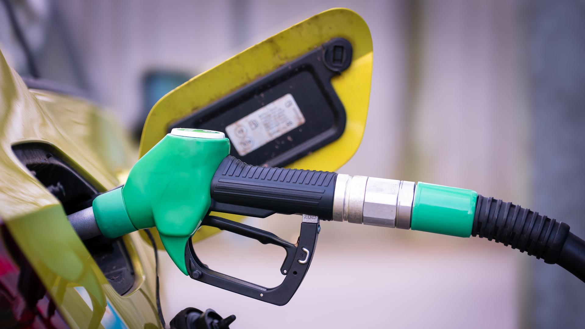 Nahaufnahme einer grünen Zapfpistole, die in den Tankstutzen eines Autos gesteckt ist. Der Tankdeckel ist geöffnet, im Hintergrund ist das Fahrzeug leicht unscharf zu sehen.