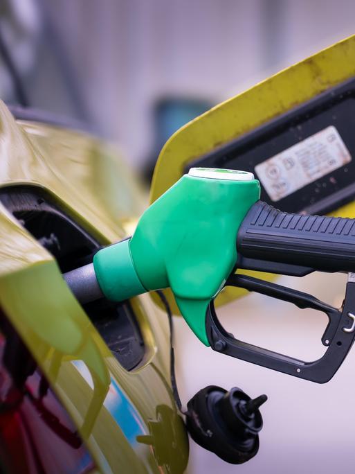 Nahaufnahme einer grünen Zapfpistole, die in den Tankstutzen eines Autos gesteckt ist. Der Tankdeckel ist geöffnet, im Hintergrund ist das Fahrzeug leicht unscharf zu sehen.