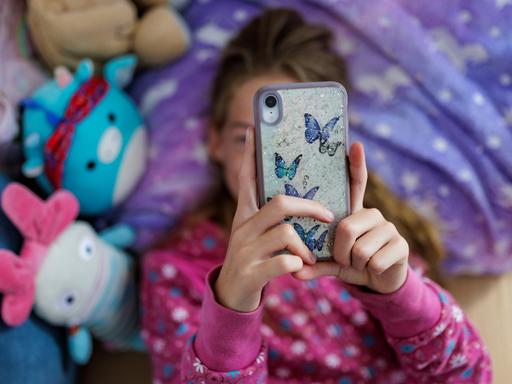 Ein Mädchen liegt in ihrem Bett und schaut auf ein Handy, das ihr Gesicht verdeckt.