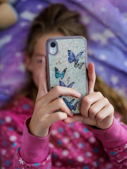 Ein Mädchen liegt in ihrem Bett und schaut auf ein Handy, das ihr Gesicht verdeckt.