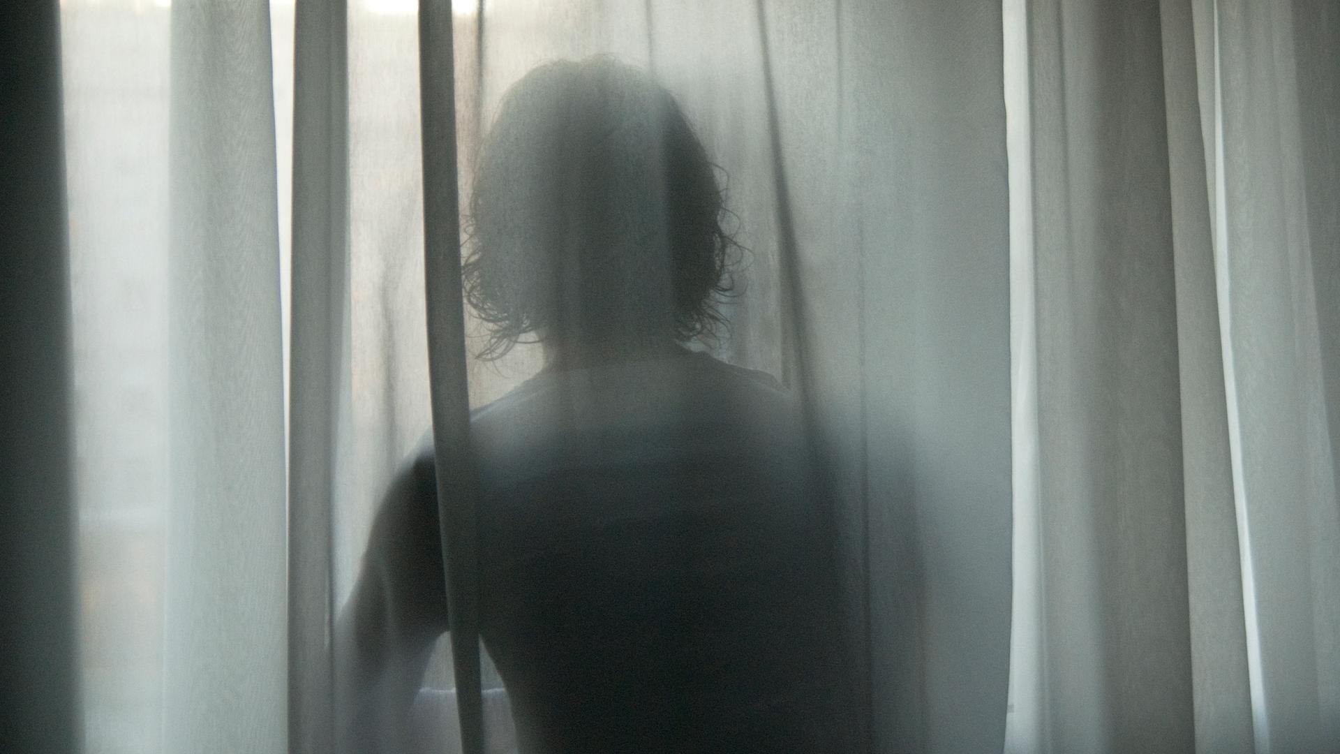 Hinter einer wehende Gardine zeichnen sich die Konturen einer Frau ab, die an einem Fenster steht. Hinter einer wehende Gardine zeichnen sich die Konturen einer Frau ab, die an einem Fenster steht.