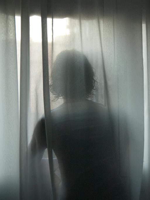 Hinter einer wehende Gardine zeichnen sich die Konturen einer Frau ab, die an einem Fenster steht. Hinter einer wehende Gardine zeichnen sich die Konturen einer Frau ab, die an einem Fenster steht.