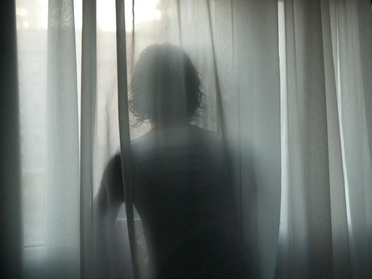Hinter einer wehende Gardine zeichnen sich die Konturen einer Frau ab, die an einem Fenster steht. Hinter einer wehende Gardine zeichnen sich die Konturen einer Frau ab, die an einem Fenster steht.