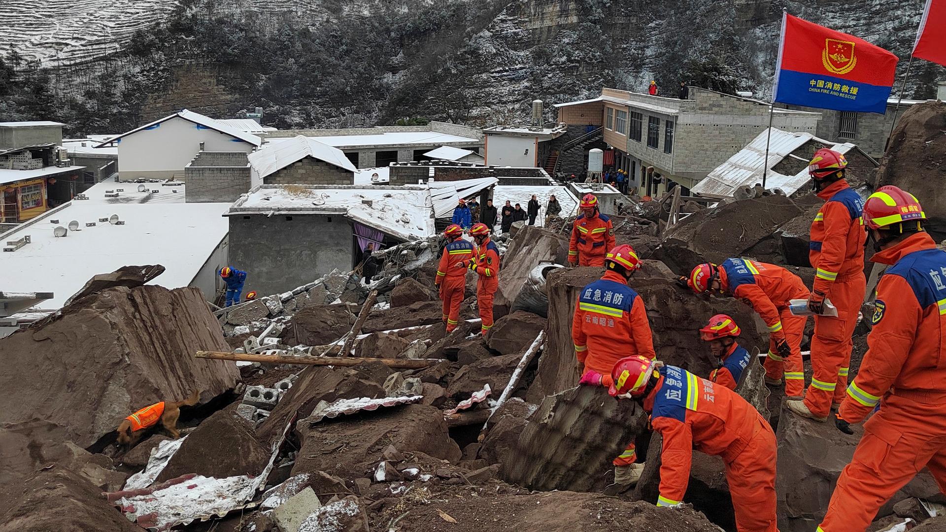 Zahlreiche Rettungskräfte in roten Overalls stehen auf einem Trümmerfeld neben zerstörten Häusern. Im Hintergrund verschneite Berge.