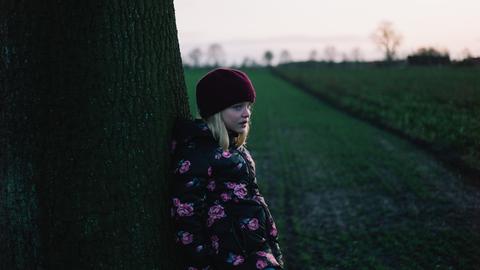Ein Kind steht auf einem Feld, den Rücken an einen Baum gelehnt.