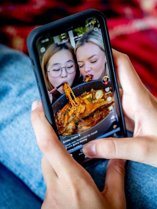 Ein Mädchen hält ein Mobiltelefon in den Händen, auf dem ein Video aus den sozialen Medien läuft. Darin filmen sich zwei Teenager beim Essen von Spaghetti.