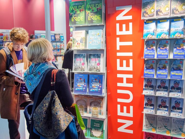 Die ersten Besucher schauen auf die Neuerscheinungen auf der Buchmesse. Sie stehen in einem großen Regal.