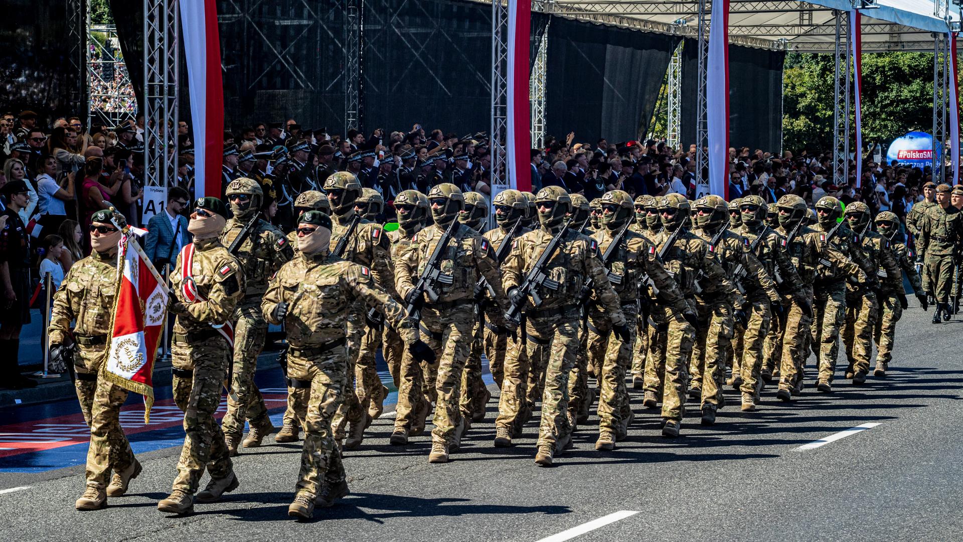 Soldaten in Tarnuniformen gehen in Reih und Glied an deiner Besuchertribüne vorbei, die mit polnischen Fahnen geschmückt ist. 