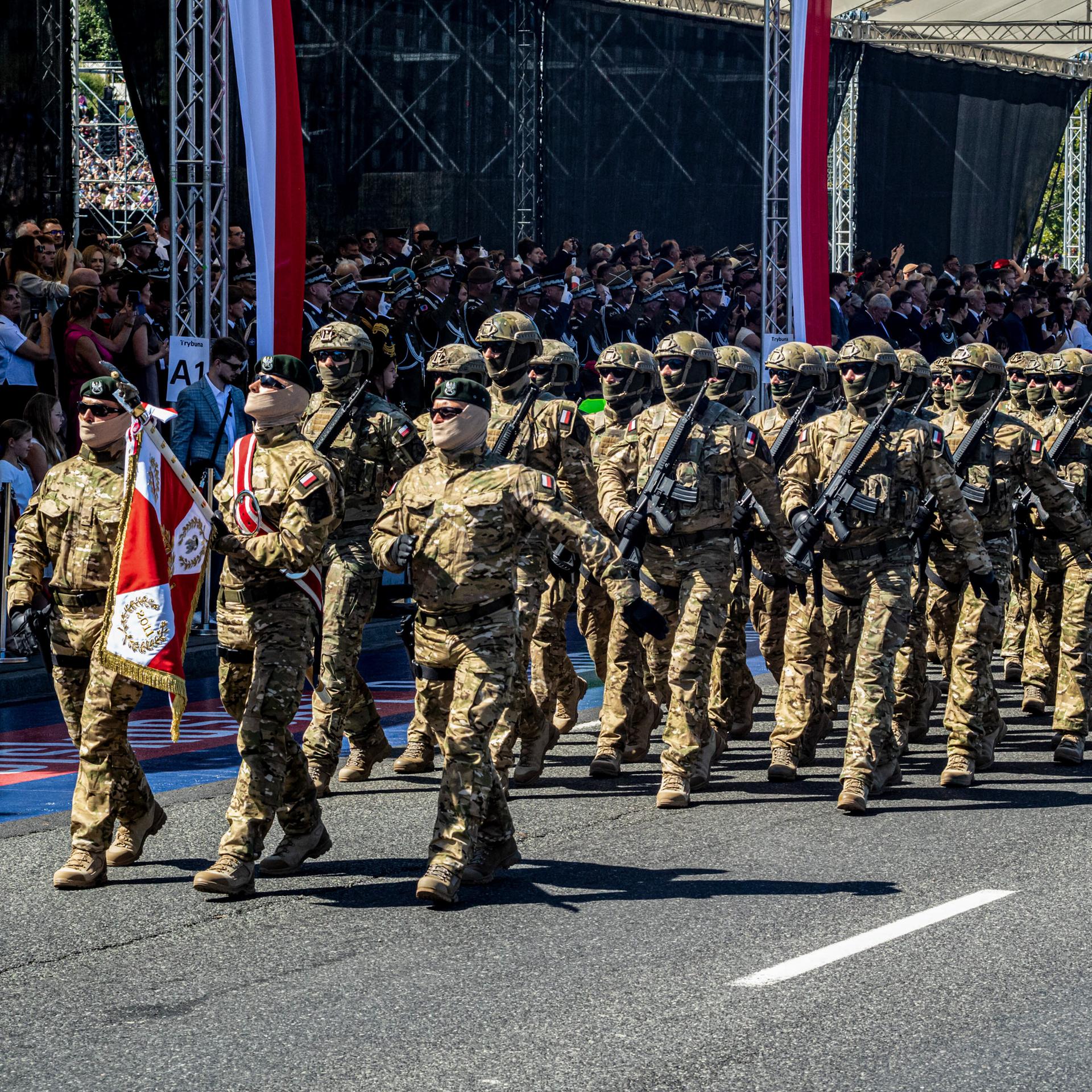 Soldaten in Tarnuniformen gehen in Reih und Glied an deiner Besuchertribüne vorbei, die mit polnischen Fahnen geschmückt ist. 