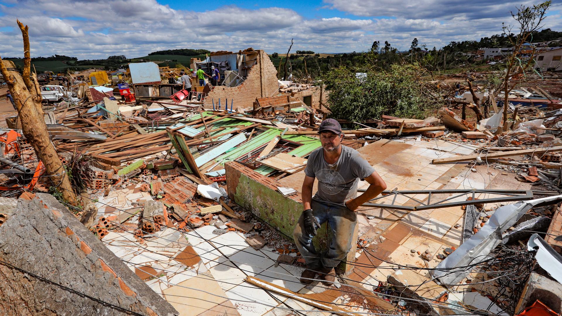 Ein Mann mit Kappe und dreckiger Kleidung sitzt auf einem Rest Mauer seines zerstörten Hauses. Um ihn herum Trümmerteile. Unter ihm ist noch ein Fliesenboden zu erkennen. Ein Mann mit Kappe und dreckiger Kleidung sitzt auf einem Rest Mauer seines zerstörten Hauses. Um ihn herum Trümmerteile. Unter ihm ist noch ein Fliesenboden zu erkennen.