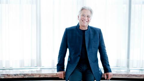 Håkan Hardenberger steht in einem dunkelblauen Anzug vor einem großen Holztisch, an den er sich lachend lehnt.
