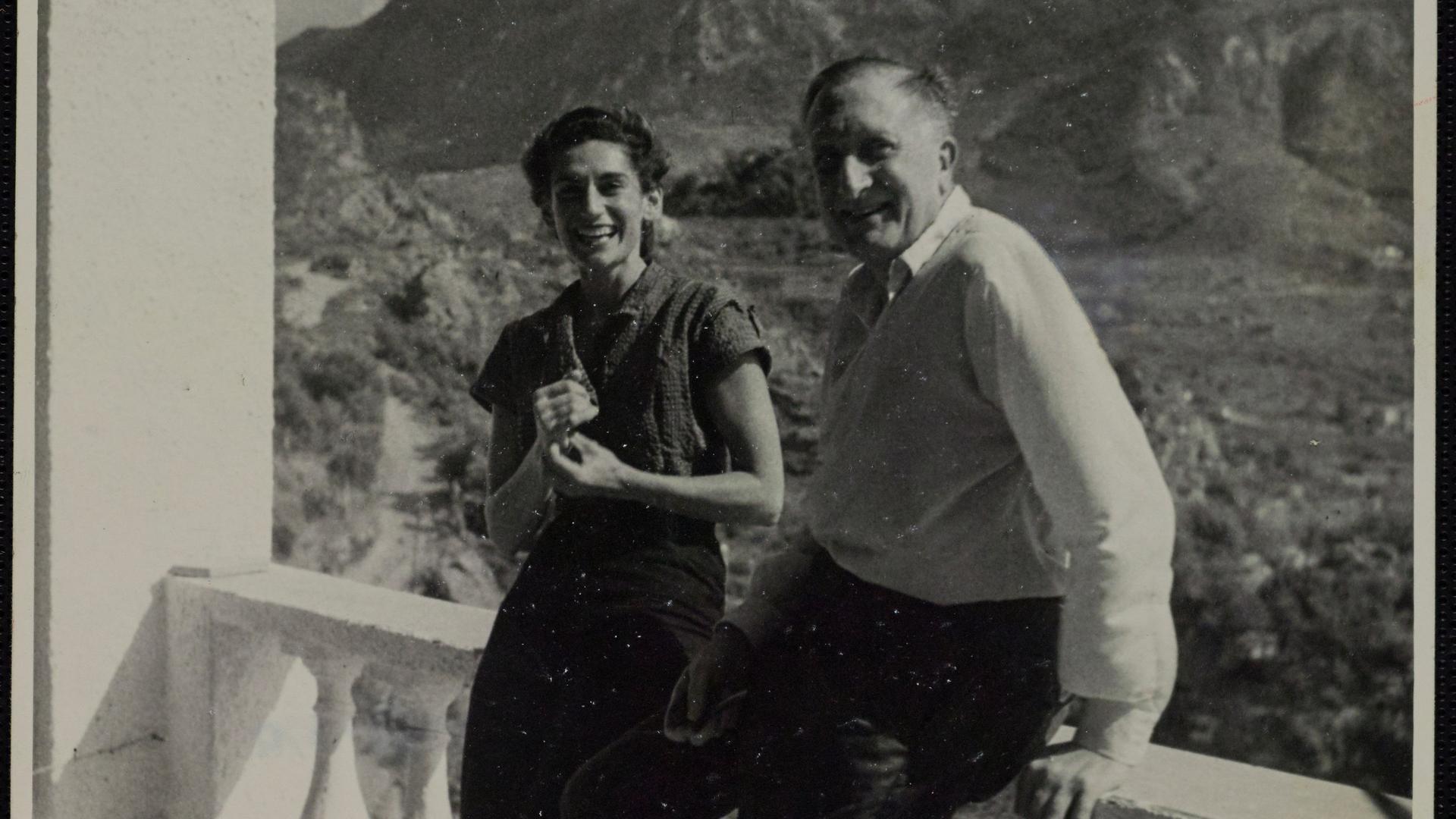 Wir sehen den Komponisten William Walton zusammen mit seiner Frau in die Kamera lächeln. Sie stehen auf einem Balkon. Hinter ihnen sieht man eine Berglandschaft. 