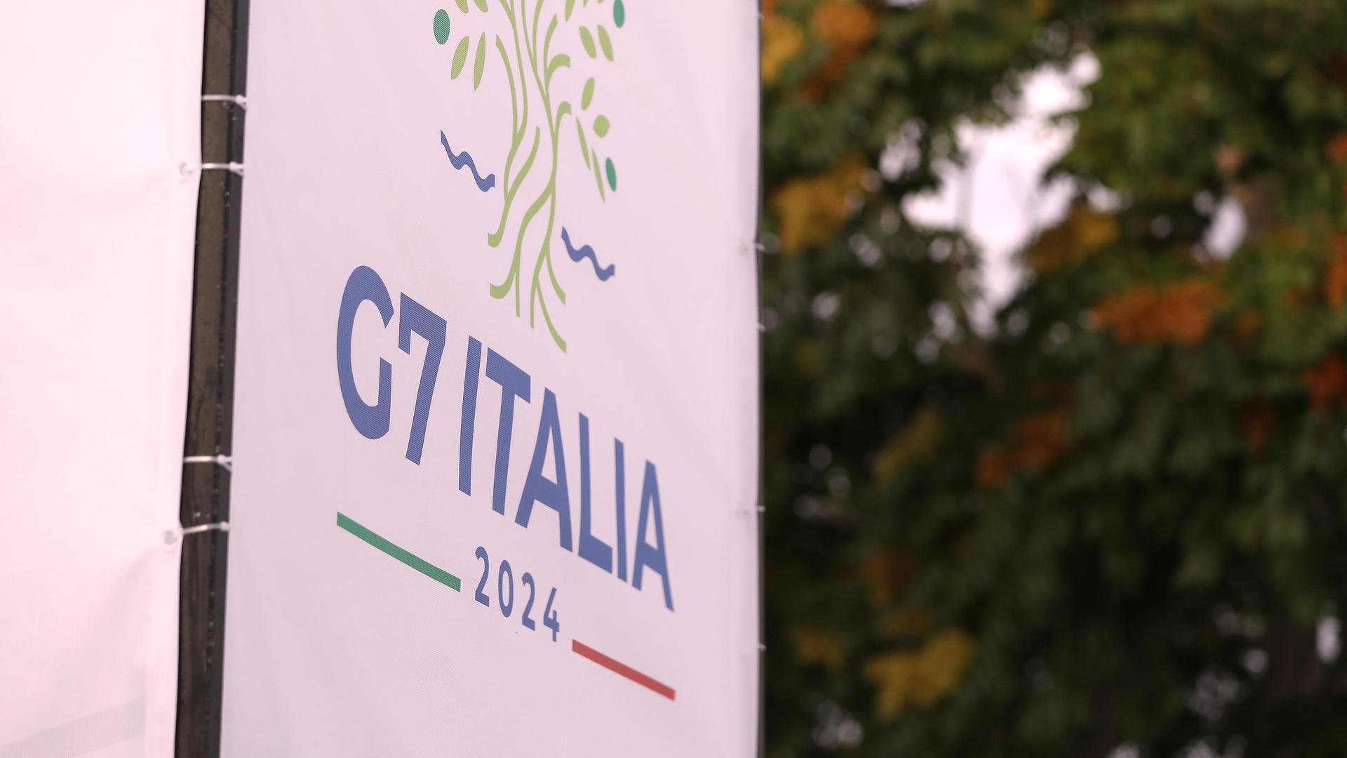 Das Logo des G7-Gipfels in Italien auf einem Banner, im Hintergrund sind Bäume zu erkennen. 