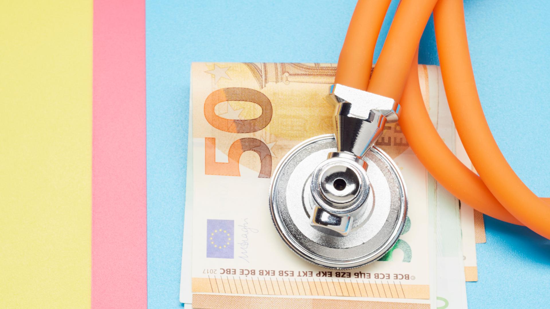 Auf einem farbigen Papieruntergrund liegen mehrere 50-Euro-Scheine. Darauf liegt ein Stethoskop.