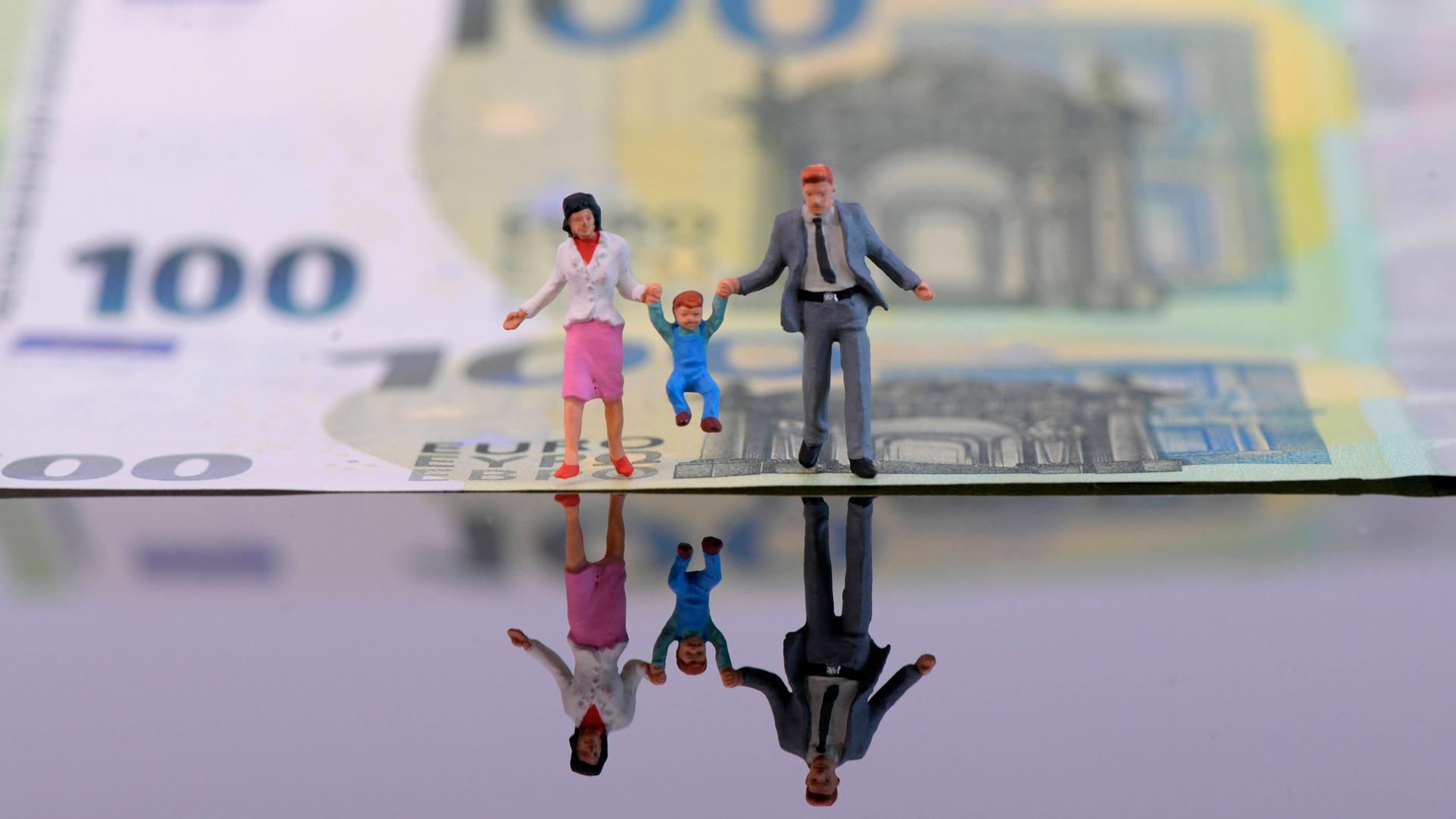 Spielfiguren von Eltern mit einem Kleinkind sind für ein Symbolfoto vor einem 100-Euro-Schein platziert. 