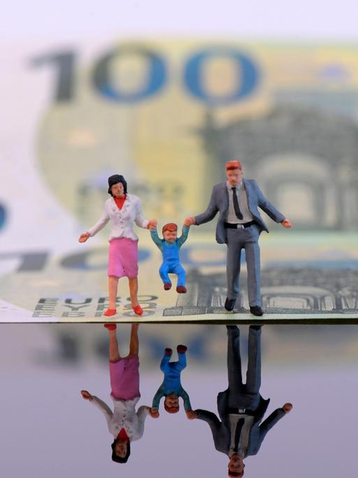 Spielfiguren von Eltern mit einem Kleinkind sind für ein Symbolfoto vor einem 100-Euro-Schein platziert. 