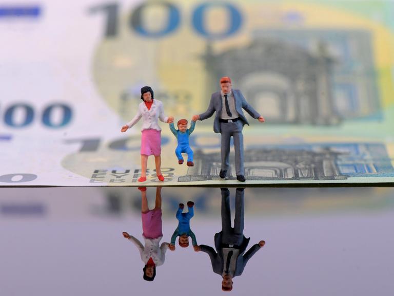 Spielfiguren von Eltern mit einem Kleinkind sind für ein Symbolfoto vor einem 100-Euro-Schein platziert. 