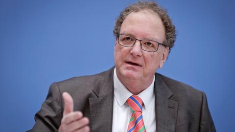 Älterer Mann mit hoher Stirm und angegrautem gelocktem Haar sitzt im Anzug vor einer blauen Wand und hebt eine Hand geradeaus vor seinen Oberkörper.