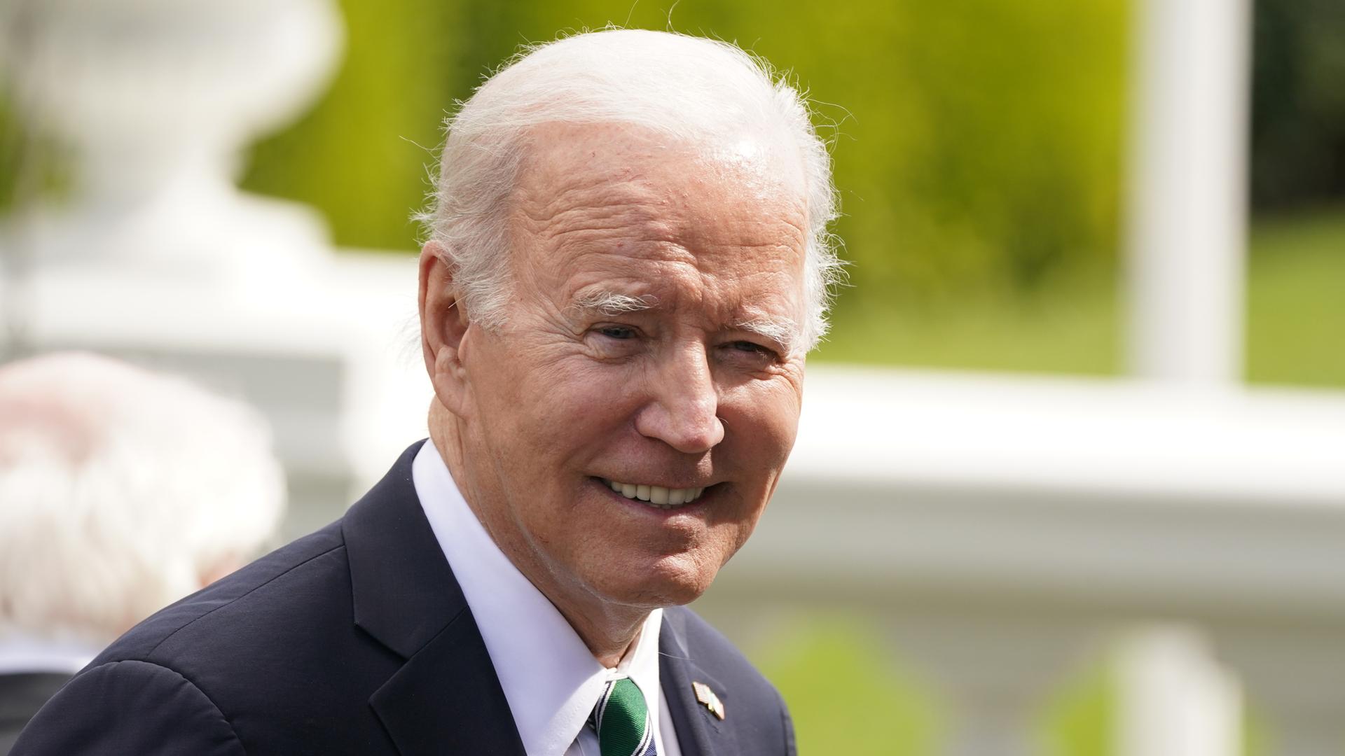 Porträtbild von US-Präsident Joe Biden: Er lächelt, im Hintergrund sind sonniges Wetter und grüne Pflanzen zu sehen.