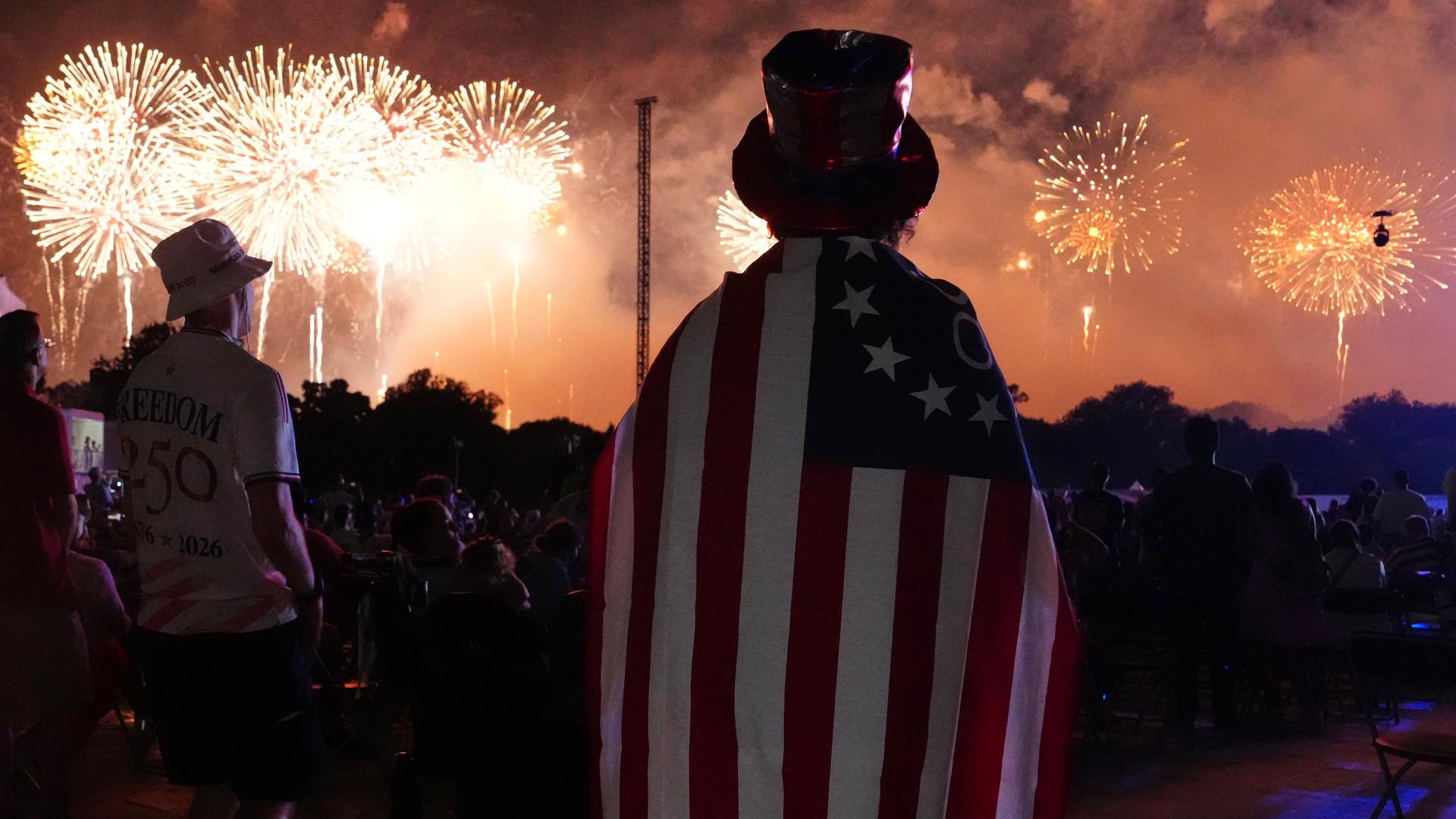 Menschen verfolgen das Feuerwerk in Washington. Sie sind von hinten zu sehen. Ein Mensch hat sich eine amerikanische Flagge umgehängt und trägt einen Zylinder. Menschen verfolgen das Feuerwerk in Washington. Sie sind von hinten zu sehen. Ein Mensch hat sich eine amerikanische Flagge umgehängt und trägt einen Zylinder.