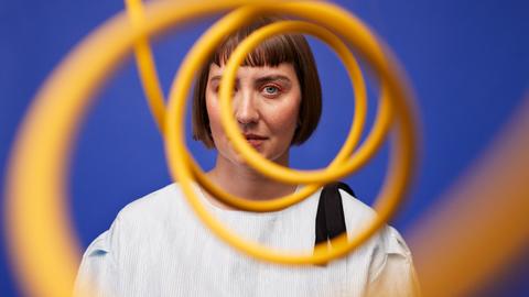 Eine Frau mit orangem Augen-Makeup steht vor einer blauen Wand und schaut durch eine gelbe Sprale, die ihr Gesicht zur Hälfte verdeckt.
