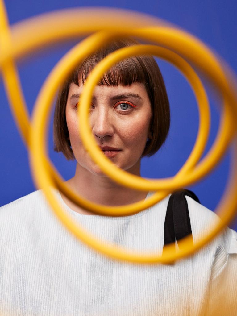 Eine Frau mit orangem Augen-Makeup steht vor einer blauen Wand und schaut durch eine gelbe Sprale, die ihr Gesicht zur Hälfte verdeckt.
