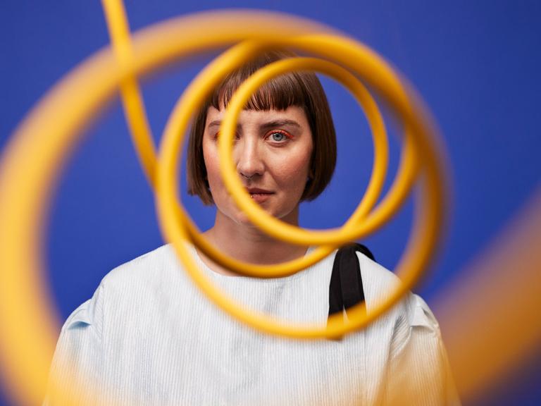 Eine Frau mit orangem Augen-Makeup steht vor einer blauen Wand und schaut durch eine gelbe Sprale, die ihr Gesicht zur Hälfte verdeckt.