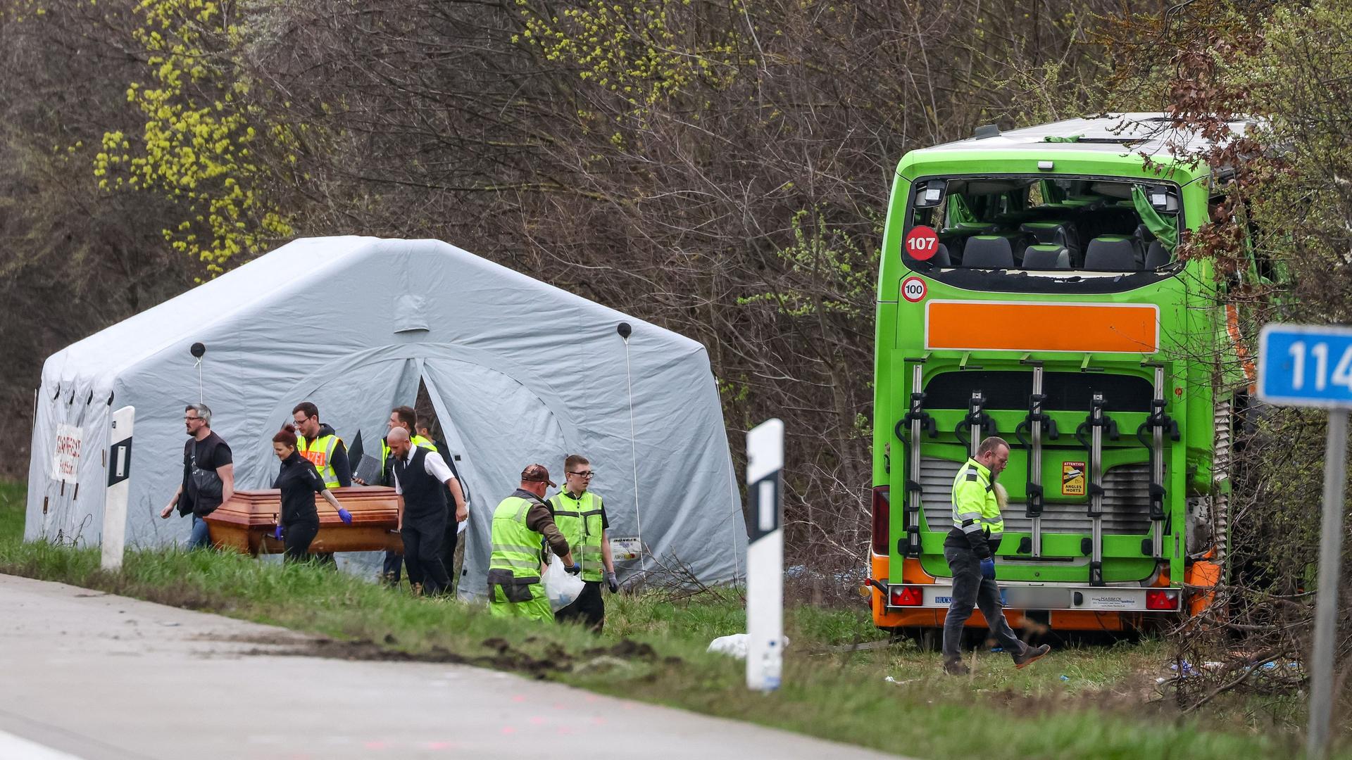 Bestatter tragen ein Todesopfer vom aufrecht stehenden  Wrack des Busses weg. Hinter ihnen ein weißes Zelt.
