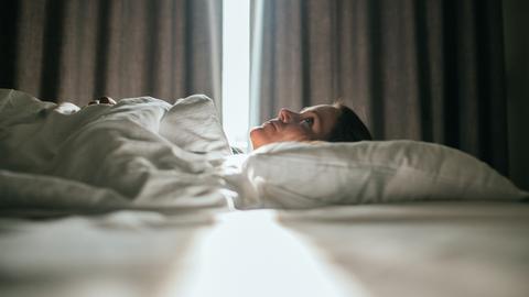 Eine Frau liegt im Bett. Sonniges Tageslicht fällt durch die Vorhänge im Hintergrund. Sie schaut zur Decke des Zimmers.