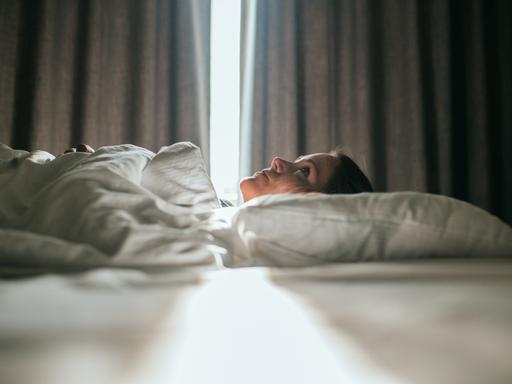 Eine Frau liegt im Bett. Sonniges Tageslicht fällt durch die Vorhänge im Hintergrund. Sie schaut zur Decke des Zimmers. Eine Frau liegt im Bett. Sonniges Tageslicht fällt durch die Vorhänge im Hintergrund. Sie schaut zur Decke des Zimmers.