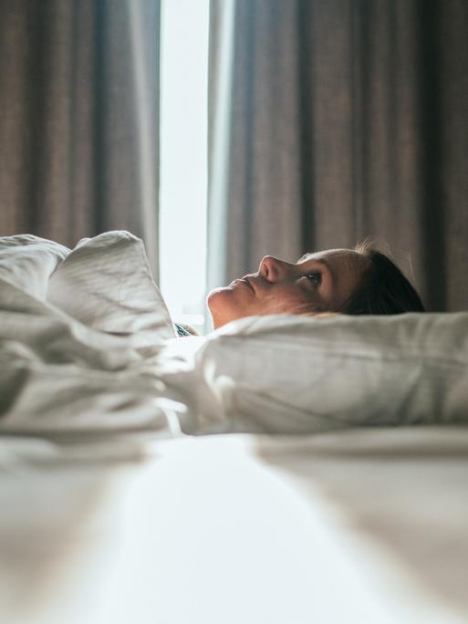 Eine Frau liegt im Bett. Sonniges Tageslicht fällt durch die Vorhänge im Hintergrund. Sie schaut zur Decke des Zimmers. Eine Frau liegt im Bett. Sonniges Tageslicht fällt durch die Vorhänge im Hintergrund. Sie schaut zur Decke des Zimmers.