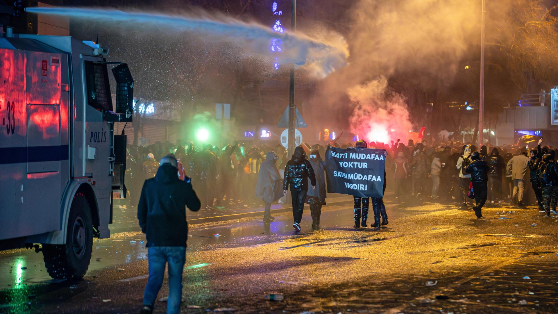 Ein Wasserwerfer zielt auf eine Gruppe demonstrierender Menschen, die ein Banner festhalten. 