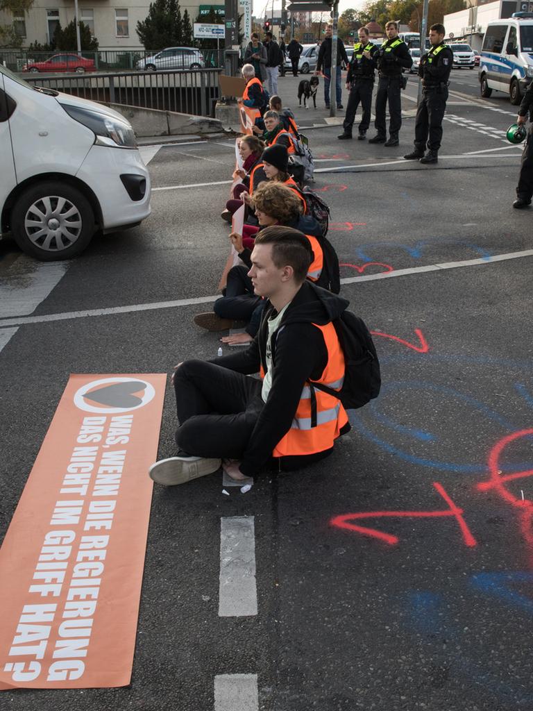 Junge Menschen in orangenen Warnwesten sitzen in einer Reihe auf einer Straße und blockieren den Autoverkehr.