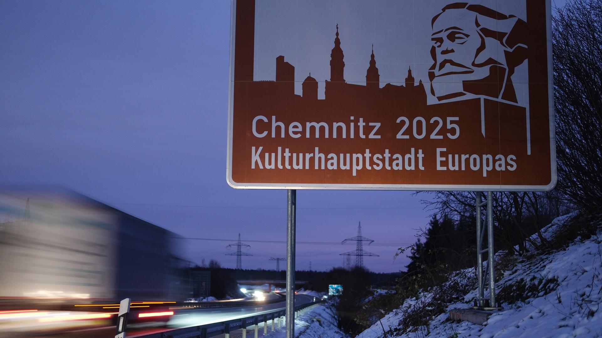 Es wird gerade dunkel. Neben dem Schild mit einer stilisierten Stadtansicht und dem Marx-Denkmal fährt ein Lastwagen vorbei 