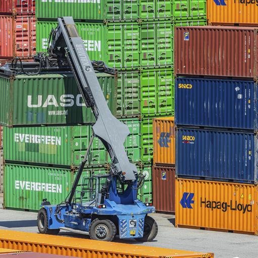 Das Bild zeigt gestapelte Frachtcontainer in einem Containerterminal. Ein Kran transportiert einen Container.
