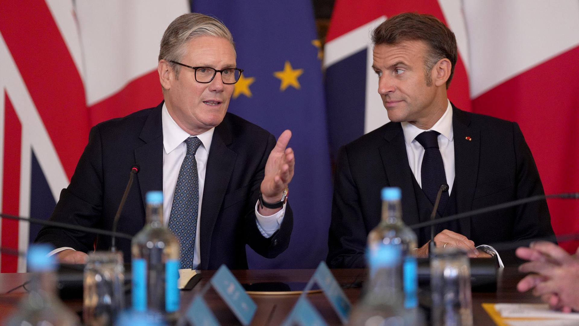 Der britische Premierminister Keir Starmer, links im Bild, sitzt neben Frankreichs Präsident Emmanuel Macron. Sie reden miteinander. Im Hintergrund stehen die britische und französische Flagge.