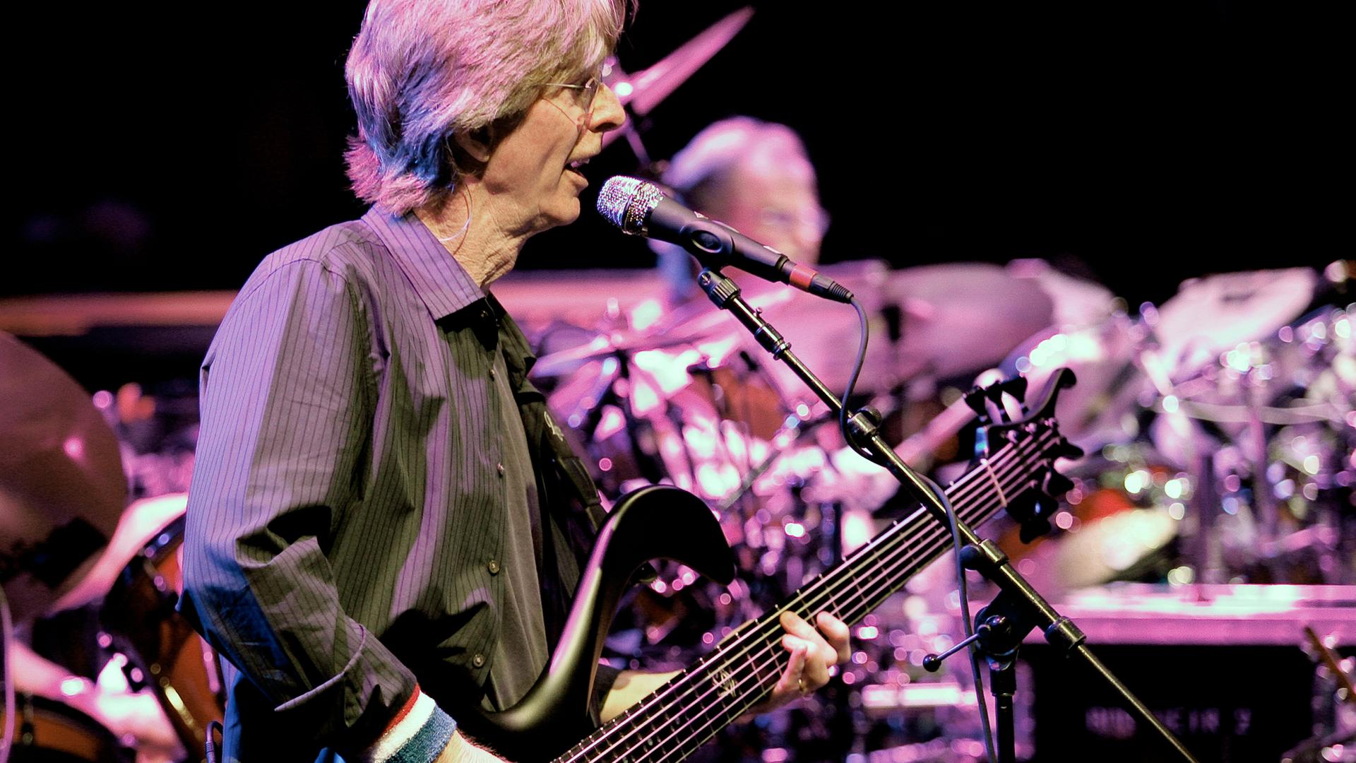 Phil Lesh, ein Mann mit längeren grauen Haaren und Brille, steht mit einer Gitarre auf der Bühne und singt in ein Mikrofon.