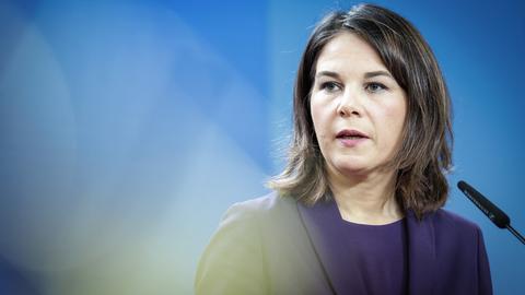 Eine junge Frau mit halblangen, dunklen Haaren wird im leicht seitlichen Profil gezeigt. Sie steht vor einem Mikrofon und trägt einen dunkelblauen Blazer, darunter ein dunkelblaues Shirt.