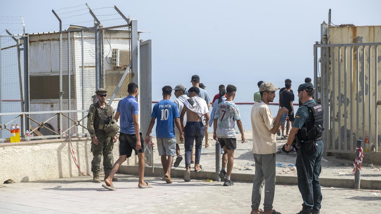 Mehrere Männer gehen durch einen abgesperrten Kontrollbereich in Ceuta. Spanische Sicherheitskräfte stehen neben ihnen. Im Hintergrund sind hohe Metallzäune und das Meer zu sehen.