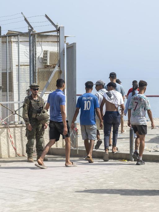 Mehrere Männer gehen durch einen abgesperrten Kontrollbereich in Ceuta. Spanische Sicherheitskräfte stehen neben ihnen. Im Hintergrund sind hohe Metallzäune und das Meer zu sehen. Mehrere Männer gehen durch einen abgesperrten Kontrollbereich in Ceuta. Spanische Sicherheitskräfte stehen neben ihnen. Im Hintergrund sind hohe Metallzäune und das Meer zu sehen.