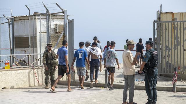 Mehrere Männer gehen durch einen abgesperrten Kontrollbereich in Ceuta. Spanische Sicherheitskräfte stehen neben ihnen. Im Hintergrund sind hohe Metallzäune und das Meer zu sehen. Mehrere Männer gehen durch einen abgesperrten Kontrollbereich in Ceuta. Spanische Sicherheitskräfte stehen neben ihnen. Im Hintergrund sind hohe Metallzäune und das Meer zu sehen.